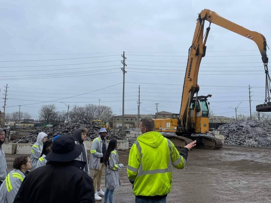 A group of students standing at a recycling center in front of heavy crane equipment and piles of metal